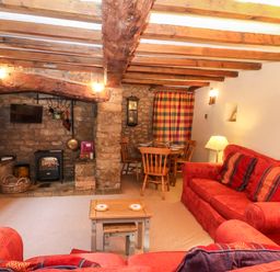 A living room with red sofas wooden beams a stone fireplace and a dining table at Spring Cottage in Wroxton