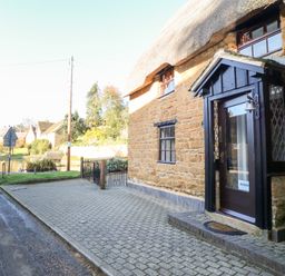 A stone cottage with a thatched roof and black porch on a street near a small body of water at Spring Cottage in Wroxton