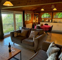 A living room with sofas wine bottle and glasses dining table kitchen area and large windows at Treetops in Woody Bay
