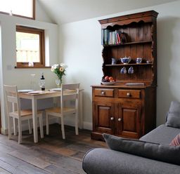 A dining area with a wooden table and two chairs next to a wooden cupboard and a gray sofa at Pops Barn in Downton