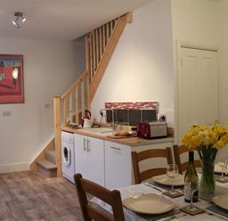 A dining table set with plates glasses and a vase of yellow flowers next to a kitchen area with white cabinets a washing machine and wooden staircase at Rojoy in Salisbury
