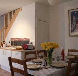 A dining table set with plates glasses and a vase of yellow flowers near a small kitchen under wooden stairs at Rojoy in Salisbury