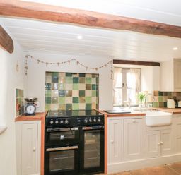 A kitchen with a black stove and oven, a white farmhouse sink, wooden countertops, cream cabinets, and green tiled backsplash at Cosy Cot in Stapleford
