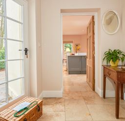 A hallway with a wicker basket on the floor a wooden table with a plant a phone and a decorative item a round mirror on the wall and a view into a kitchen at East Lodge in Rockbourne