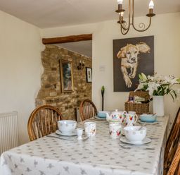 A dining room with a set table featuring bowls cups and a teapot with dog and rooster artwork on the walls at Henmarsh Cottage in Moreton-In-Marsh