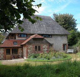 A thatched roof house with brick and stone walls surrounded by trees and a garden with a white bench at The Yeoman's House in Bignor