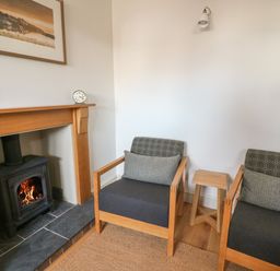 A room with two wooden armchairs with cushions a small wooden stool a wood burning stove in a wooden fireplace and a framed picture on the wall at Craigluscar in Newport Pembrokeshire