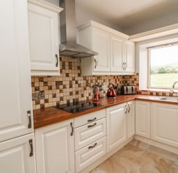 A kitchen with white cabinets brown tiled backsplash wooden countertops a stovetop a stainless steel range hood a red kettle a toaster a window over the sink showing a grassy landscape at Dromstabla in Glencar