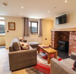 A living room with grey sofas a wooden coffee table a brick fireplace and a wall-mounted TV at Llety Dewi in Lake Vyrnwy