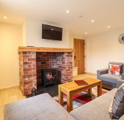 A living room with a brick fireplace and wood stove a wall-mounted TV grey sofas a wooden coffee table and a clock on the wall at Llety Dewi in Lake Vyrnwy