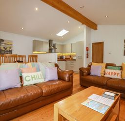 A living room with brown leather sofas and colorful cushions next to a wooden dining table and an open kitchen at 6 Streamside in Lanreath