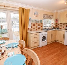A kitchen with wooden cabinets a washing machine a fridge a dining table set with blue plates and glasses and yellow curtains at The Chace in Portreath