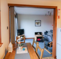A dining area with a wooden table and chairs leading into a living room with a gray sofa a wooden coffee table a TV and wall art at Kestrel Lodge in Dumfries