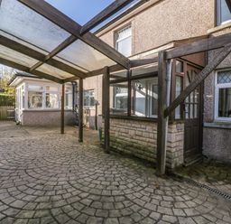 An outdoor patio area with patterned paving and a wooden pergola attached to a house with pebble dash walls and white framed windows at Rose Cottage in Bream
