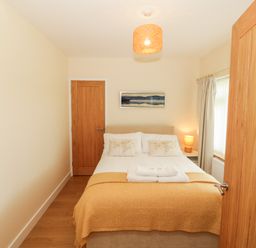 A bedroom with a double bed, bedside table with lamp, wooden doors, and a window with curtains at Llain Bach in Llangefni