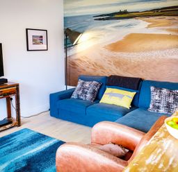 A living room with a blue sofa a wooden TV stand a brown chair and a bowl of fruit at Oystercatcher in Embleton