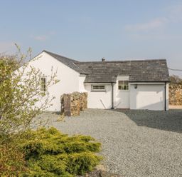 A white cottage with a slate roof and gravel driveway surrounded by bushes at Rhos Y Foel Cottage in Nefyn