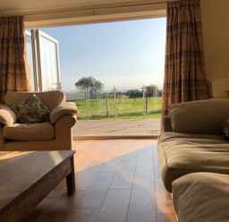 A living room with beige armchairs, a wooden coffee table, curtains, and an open glass door showing a green field outside at Rhos Y Foel Cottage in Nefyn
