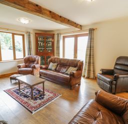 A living room with leather sofas a wooden coffee table a rug a wooden cabinet and windows with curtains at Pentrego Isaf in Meifod