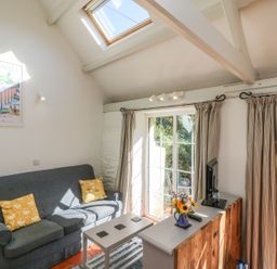 A living room with a blue sofa with yellow cushions a small white coffee table a TV on a wooden cabinet and glass doors with curtains at The Daffodil Shed in Bere Alston