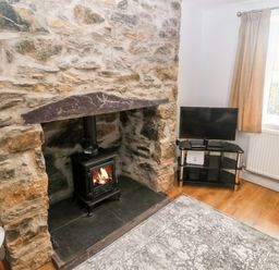 A room with a stone fireplace and wood burning stove and a television on a stand next to a window at 3 Green Terrace in Llangaffo near Newborough