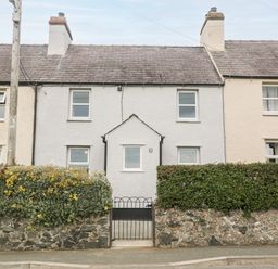 A stone cottage with white walls and two chimneys behind a stone wall and garden hedge at 3 Green Terrace in Llangaffo near Newborough