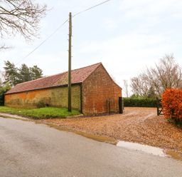 A brick barn with a tiled roof next to a gravel driveway and trees at Honey Buzzard Barn in Fakenham