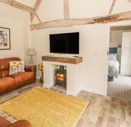 A living room with brown leather sofas a yellow rug a wood stove under a wall mounted TV and a doorway leading to a bedroom at Megs Cottage in Tiddington