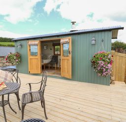 A wooden deck with metal table and chairs outside a green cabin with open wooden doors and hanging flower baskets at Ivys Way in Rhayader
