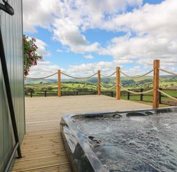 An outdoor deck with a hot tub and rope railing overlooking green hills and a cloudy sky at Ivys Way in Rhayader