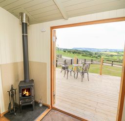 A room with a wood burning stove and a view of a wooden deck with a table and two chairs overlooking green fields at Ivys Way in Rhayader