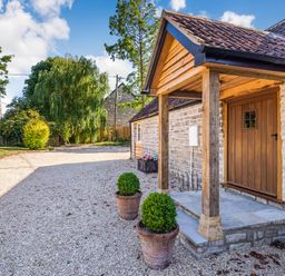 The entrance of a stone house with a wooden door covered by a porch and gravel driveway with potted plants at Old Farm Stables South Barrow near Sparkford