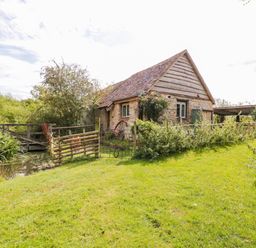 A stone and wood house with a pitched roof next to a wooden gate and a pond in a grassy area at Jackdaw's Roost in Cheltenham