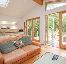 A living room with a leather sofa and cushions near glass doors opening to a deck with wicker chairs at 4 Stonerush Valley in Lanreath