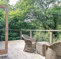 A wooden deck with two wicker chairs and a wicker table surrounded by trees at 4 Stonerush Valley in Lanreath