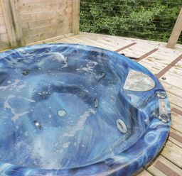 A blue outdoor hot tub on a wooden deck with railing and green trees in the background at 7 Streamside Lanreath