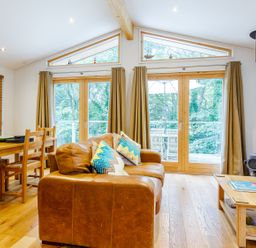 A living room with a brown leather sofa colorful pillows a wooden coffee table dining table with chairs large windows and a wood stove at 8 Streamside in Lanreath