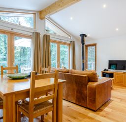 A living room with a wooden dining table and chairs a brown leather sofa a TV on a wooden stand and large windows with curtains at 8 Streamside in Lanreath