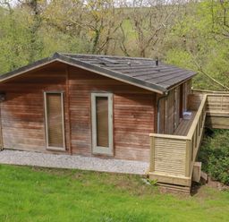 A wooden cabin with a pitched roof and a deck surrounded by trees and grass at 2 Stonerush Valley in Lanreath