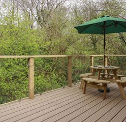 A wooden deck with a round picnic table and green umbrella surrounded by trees at 2 Stonerush Valley in Lanreath