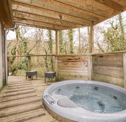 A wooden deck with a hot tub and two stools surrounded by trees at 9 Stonerush Valley in Lanreath