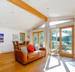 A living room with a leather sofa and wooden dining table next to large glass doors opening to a patio at 2 Water's Edge Lanreath