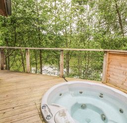 An outdoor wooden deck with a jacuzzi and a railing overlooking trees and a body of water at 2 Water's Edge in Lanreath