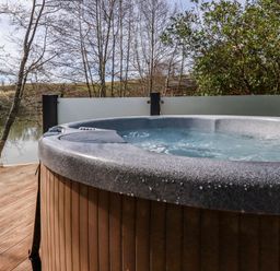 An outdoor hot tub on a wooden deck with trees and a body of water in the background at 4 Water's Edge Lanreath