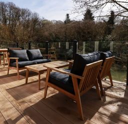 An outdoor wooden deck with wooden chairs with black cushions and tables surrounded by trees at 4 Waters Edge in Lanreath