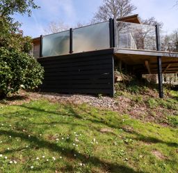 An outdoor raised deck with glass railings and black wooden panels surrounded by grass and bushes at 4 Water's Edge in Lanreath