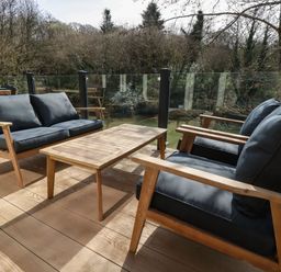 An outdoor patio with wooden furniture including a sofa, chairs, and a table with glass railing and trees in the background at 4 Water's Edge in Lanreath