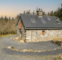 A stone cottage with skylights and wooden shutters surrounded by trees with outdoor wooden benches at Brithgwm Canol in Brithdir near Dolgellau