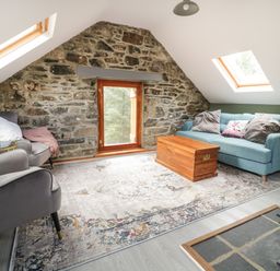 A living room with a stone wall a wooden window two skylights two armchairs a sofa a wooden chest and a rug at Brithgwm Canol in Brithdir near Dolgellau