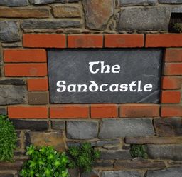 A stone wall with a sign reading The Sandcastle surrounded by red bricks and small green plants at The Sandcastle in Freshwater East